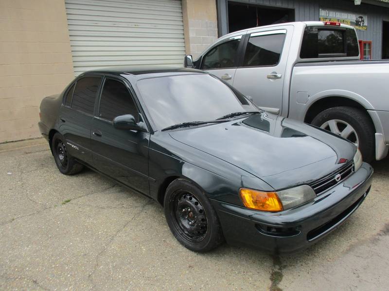 Dark green Toyota Corolla in front of a roll-up door, a silver Nissan pickup behind it and a black shop sign on the wall