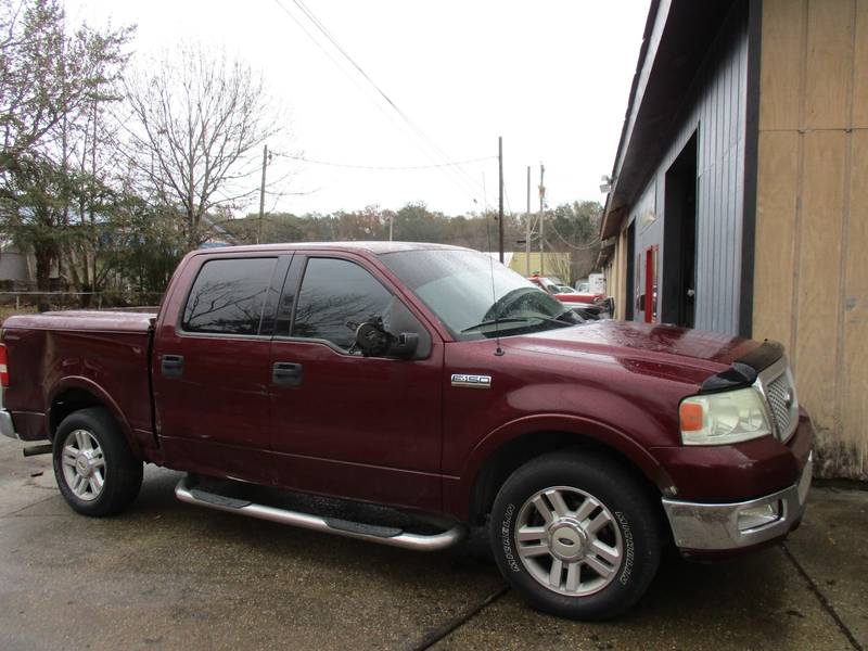 Maroon Ford F-150 crew-cab pickup with running boards, parked beside a gray metal shop building