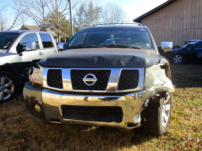 Black Nissan Titan pickup with a crushed front bumper and fender on the driver side, parked on grass by a metal shop building