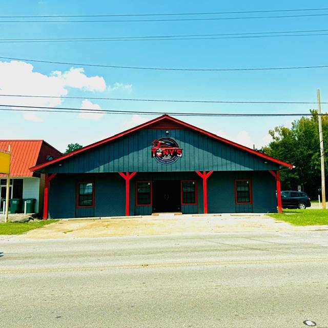 Teal-blue board-and-batten shop building with red trim and red posts, a round sign on the gable, and a concrete apron by the road