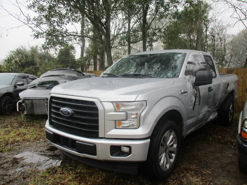 Silver Ford F-150 with a shattered windshield and a caved-in driver door, on wet ground beside other wrecked cars