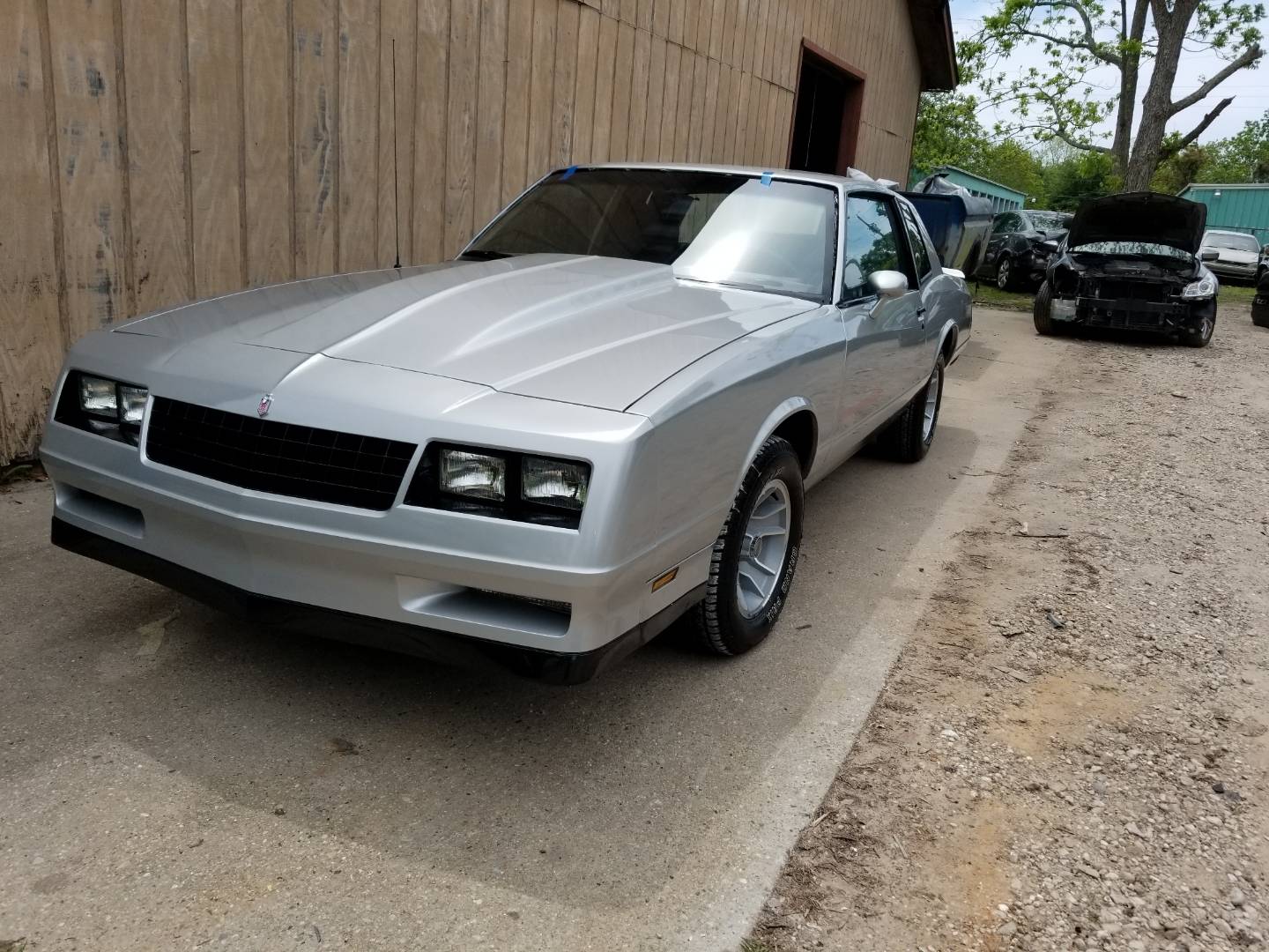 Silver 1980s coupe with glossy paint parked on concrete beside a tan wooden building, a damaged black car with its hood up and other cars behind