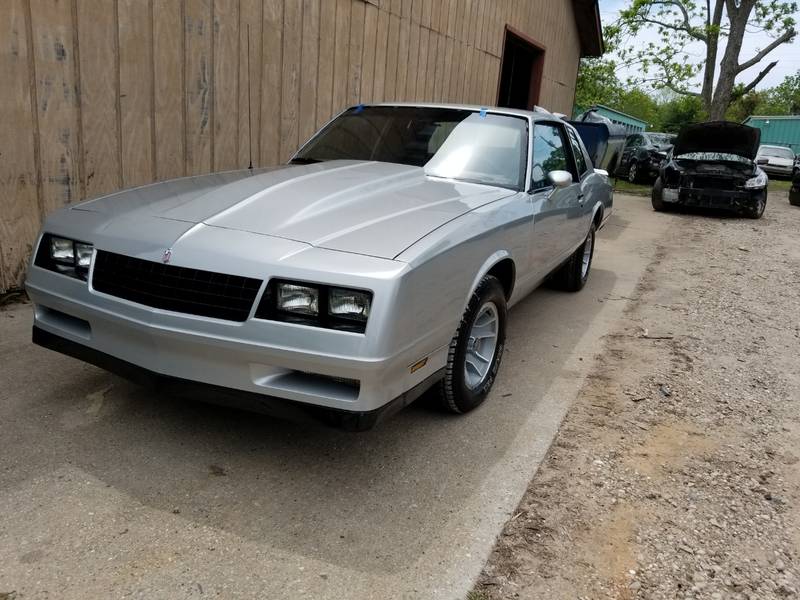 Silver 1980s coupe with glossy paint, parked along a wooden building with other cars behind